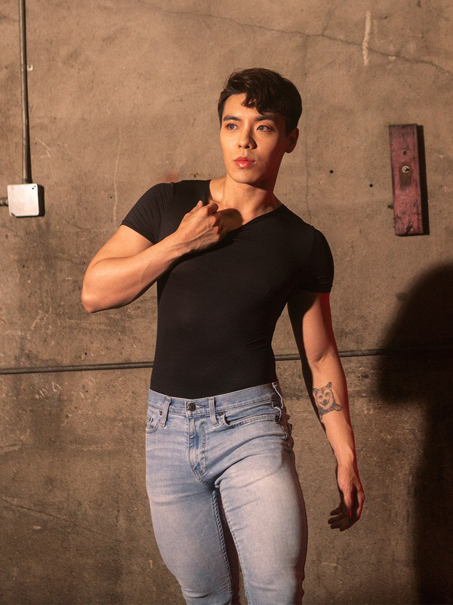 A male model wearing a black bodysuit with short-sleeves over blue jeans.
