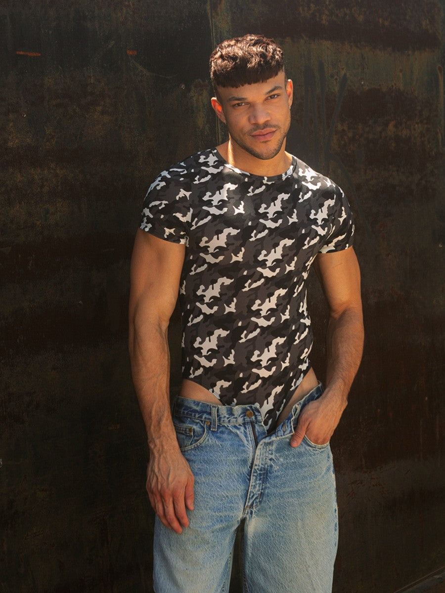 A male model  wearing a black & white camouflage bodysuit with short-sleeves over blue jeans. 