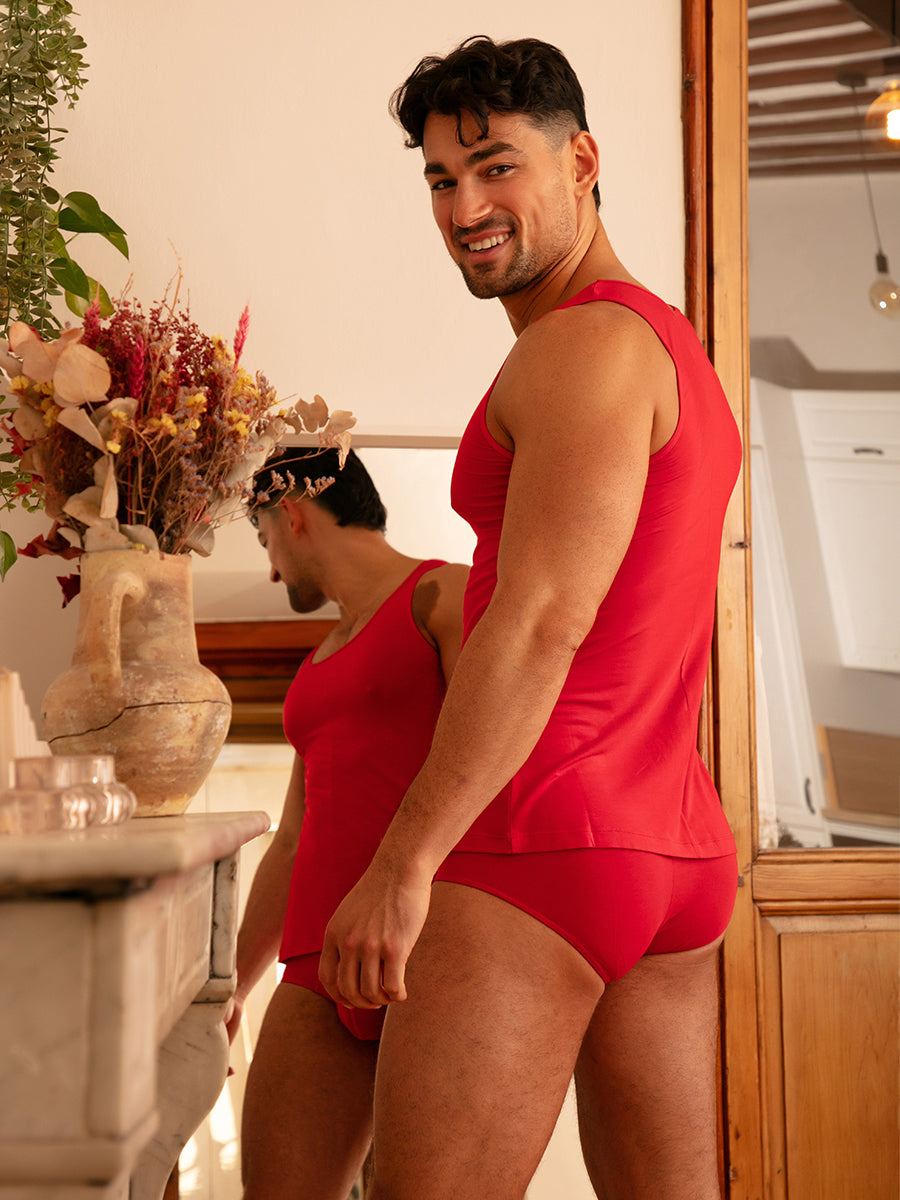 Back view of a man wearing a red tank top and matching Body Aware briefs, standing in front of a mirror.