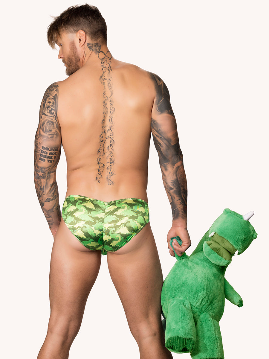 Back view of a male model wearing a green satin Body Aware brief with camo pattern holding a dinosaur plushie