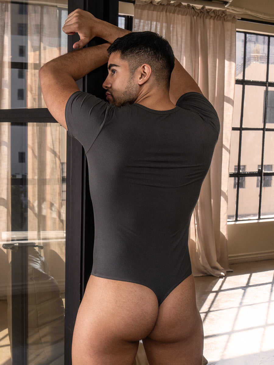 Back view of a man wearing a dark grey thong bodysuit standing next to a window.