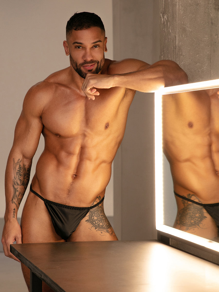 Front view of a man wearing a black satin thong, standing next to a mirror.